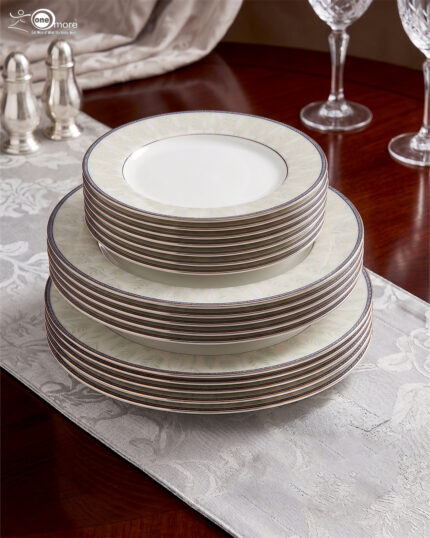 A professionally styled stack of the OneMore 18-piece super bone china dinnerware set, showcasing six quarter plates on top, six deep plates in the middle, and six large rice plates at the bottom, featuring an elegant light-green patterned border on a silver table runner