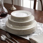 A stacked 18-piece OneMore super bone china dinnerware plate set featuring elegant cream plates with intricate lace damask borders, displayed on a dark wooden table with a silver runner and stainless steel cutlery