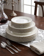 A stacked 18-piece OneMore super bone china dinnerware plate set featuring elegant cream plates with intricate lace damask borders, displayed on a dark wooden table with a silver runner and stainless steel cutlery