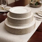 A professional shot of an 18-piece super bone china plate set stacked in three tiers, featuring elegant gold rims and subtle beige floral patterns on a luxury wooden dining table with a linen runner.