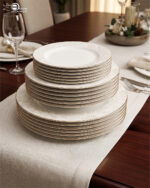 A professional shot of an 18-piece super bone china plate set stacked in three tiers, featuring elegant gold rims and subtle beige floral patterns on a luxury wooden dining table with a linen runner.