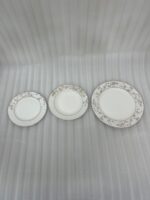 A stacked 18-piece premium super bone china dinnerware set with elegant floral and striped rim borders, neatly arranged on a dark wooden dining table next to a crystal glass and fabric napkin