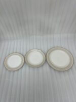 A neatly stacked 18-piece super bone china dinner plate set with elegant gold borders, arranged on a beige placemat next to a gold fork, a gold knife, and a crystal wine glass on a wooden table.