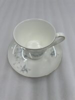 A person holding a white bone china teacup filled with milk tea, featuring a grey floral design and gold rim, resting on a matching saucer with a silver spoon on a wooden table.