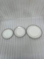 A stacked 18-piece white super bone china plate set with elegant silver-grey floral borders, displayed on a dark wooden table next to a folded cloth napkin with a knife and fork