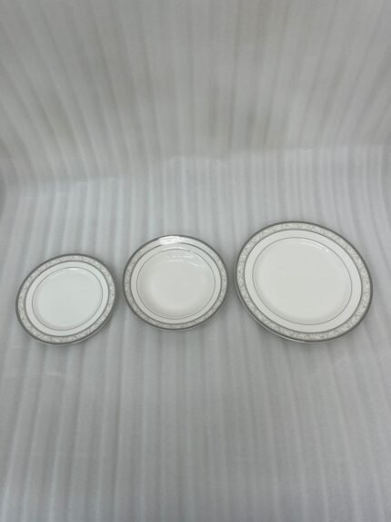 A stacked 18-piece white super bone china plate set with elegant silver-grey floral borders, displayed on a dark wooden table next to a folded cloth napkin with a knife and fork