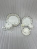 A complete 24-piece luxury tea set on a wooden table, featuring a white porcelain teapot, milk jug, sugar pot, cups, saucers, and plates with delicate silver fern patterns.