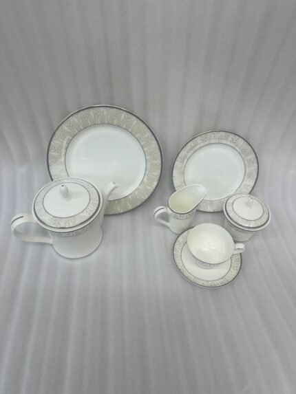 A complete 24-piece luxury tea set on a wooden table, featuring a white porcelain teapot, milk jug, sugar pot, cups, saucers, and plates with delicate silver fern patterns.