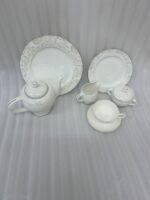 A 24-piece white porcelain tea set featuring a delicate silver floral scroll pattern, including a teapot, cups, saucers, plates, and serving pots, arranged on a wooden table.
