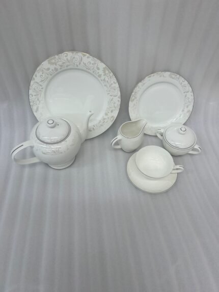 A 24-piece white porcelain tea set featuring a delicate silver floral scroll pattern, including a teapot, cups, saucers, plates, and serving pots, arranged on a wooden table.