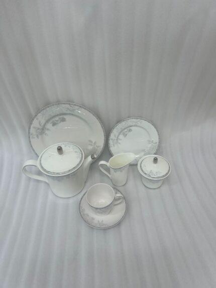 A sophisticated 24-piece white porcelain tea set featuring a delicate grey floral pattern and silver trim, arranged elegantly on a table runner.