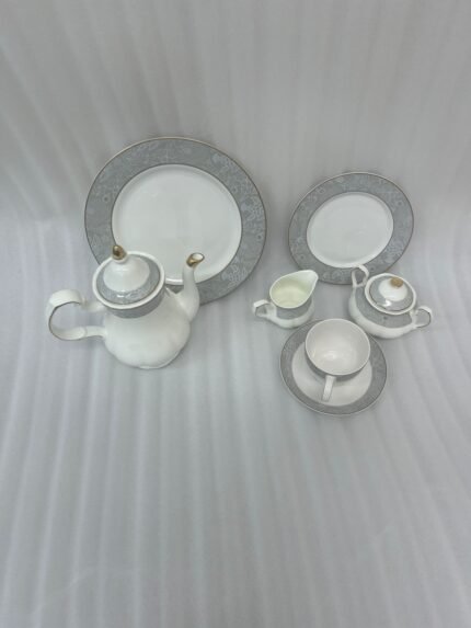 : A complete 24-piece white porcelain tea set featuring delicate grey floral patterns with gold trim, including a teapot, teacups, saucers, and plates, arranged on a wooden surface.