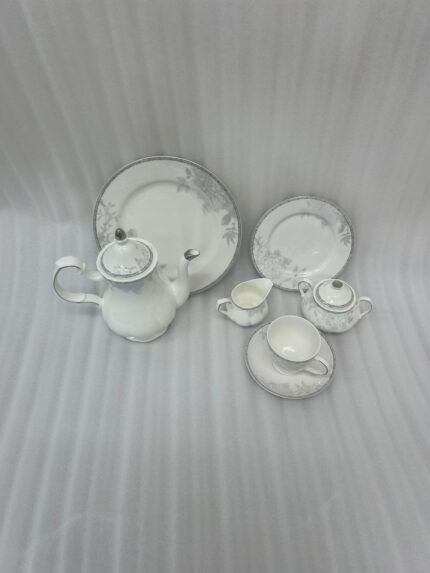 A 24-piece white porcelain tea set featuring delicate gray floral patterns and silver trim, arranged on a light wooden surface.
