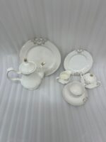 A luxurious 24-piece white porcelain tea set featuring delicate gold filigree patterns, including a teapot, teacups, saucers, plates, and serving pieces displayed on a marble-textured surface