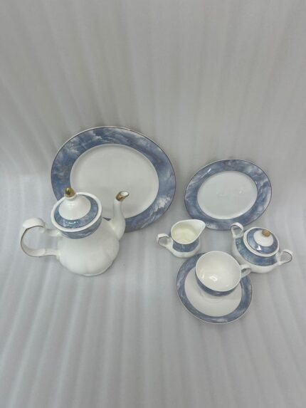 A 24-piece luxury porcelain tea set featuring a white base with elegant blue marble-patterned rims and gold accents, displayed on a textured stone surface including a teapot, cups, saucers, plates, sugar pot, and creamer.