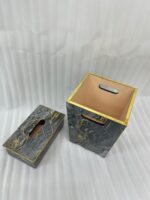 A close-up photograph of a hand pulling a tissue from a luxury black marble and gold-veined tissue box cover with gold trim, next to a matching small open-top waste bin with a warm terracotta interior and cutout handles, all on a wooden surface in a contemporary bathroom setting.