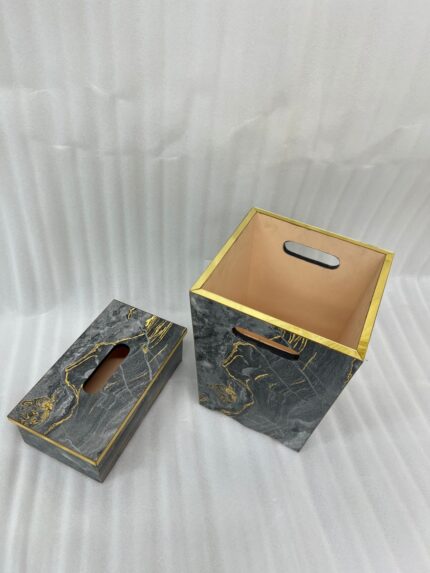 A close-up photograph of a hand pulling a tissue from a luxury black marble and gold-veined tissue box cover with gold trim, next to a matching small open-top waste bin with a warm terracotta interior and cutout handles, all on a wooden surface in a contemporary bathroom setting.