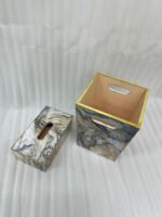 A hand dropping a tissue into a modern marble-patterned dustbin with gold trim, sitting next to a matching tissue box cover on a wooden counter.