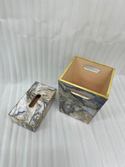 A hand dropping a tissue into a modern marble-patterned dustbin with gold trim, sitting next to a matching tissue box cover on a wooden counter.