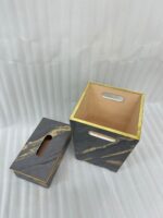 A hand with a gold ring is seen dropping a crumpled tissue into a sophisticated dark marble waste bin with prominent gold veining and trim. In the foreground is a matching tissue box cover on a wooden desk. A stacked notebook and leather portfolio are also visible.