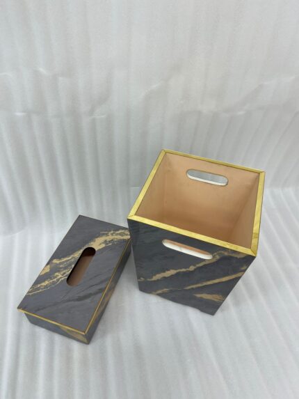 A hand with a gold ring is seen dropping a crumpled tissue into a sophisticated dark marble waste bin with prominent gold veining and trim. In the foreground is a matching tissue box cover on a wooden desk. A stacked notebook and leather portfolio are also visible.