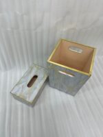A close-up view of a luxury faux-marble waste bin with intricate gold veining and polished gold trim, positioned next to a matching marble tissue box holder on a wooden desk. A hand is seen depositing a tissue into the bin, which features a peach-pink interior lining.