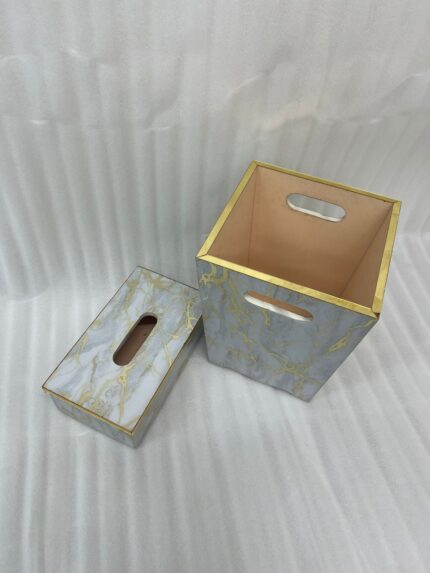 A close-up view of a luxury faux-marble waste bin with intricate gold veining and polished gold trim, positioned next to a matching marble tissue box holder on a wooden desk. A hand is seen depositing a tissue into the bin, which features a peach-pink interior lining.