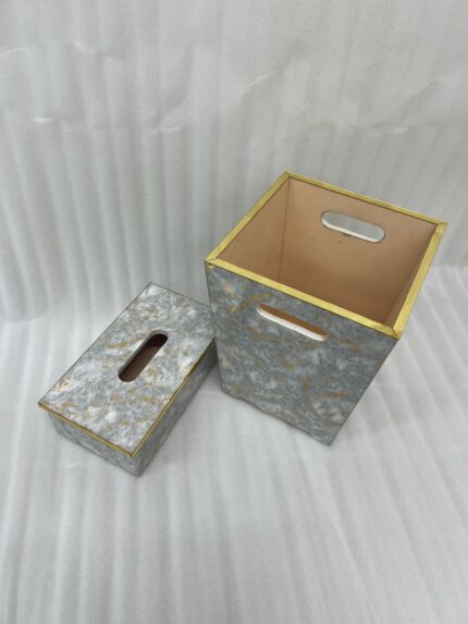 A close-up view of a hand reaching to pull a tissue from a grey and gold-veined faux-marble tissue box cover with polished gold trim, next to a matching compact grey marble waste bin with handle cutouts and a peach-colored interior, sitting on a wooden desk.