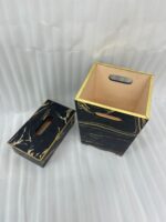 Luxury black and gold marble pattern dustbin and matching tissue box holder on a wooden vanity next to a marble bathroom sink.