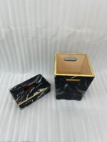 A matching executive desk accessory set featuring a black marble pattern wastebasket and a rectangular tissue box cover with elegant gold trim, placed on a dark wooden office desk.