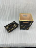 A hand pulling a tissue from a luxury black and gold marble tissue box cover next to a matching wastebasket on a table.