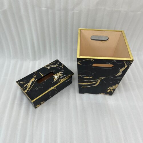 A hand pulling a tissue from a luxury black and gold marble tissue box cover next to a matching wastebasket on a table.