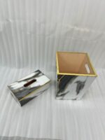 A luxury white and black marble-patterned tissue box holder and matching wastebasket with elegant gold trim, displayed on a wooden table.