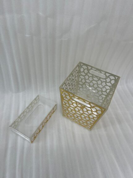 Clear acrylic tissue box holder and matching small desktop dustbin with an elegant gold geometric lattice pattern on a wooden desk.