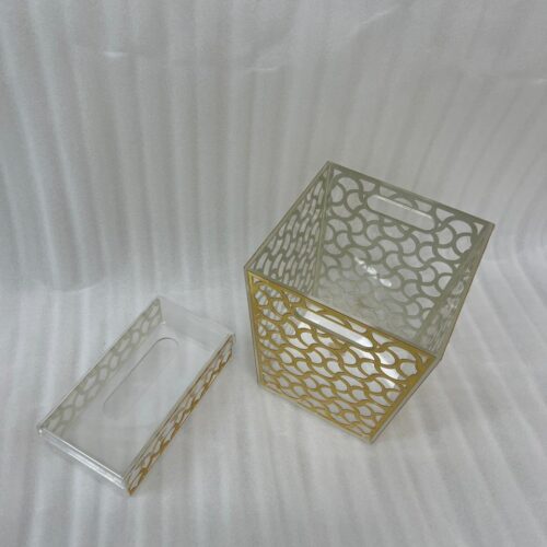 Clear acrylic tissue box holder and matching small desktop dustbin with an elegant gold geometric lattice pattern on a wooden desk.