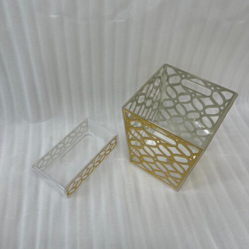 Clear acrylic dustbin and matching tissue box holder with a gold geometric lattice pattern on a white marble tabletop.