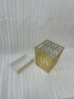An elegant gold and clear acrylic dust bin with a laser-cut leaf pattern on a wooden table, featuring a matching tissue box holder and a hand dropping a crumpled tissue inside.