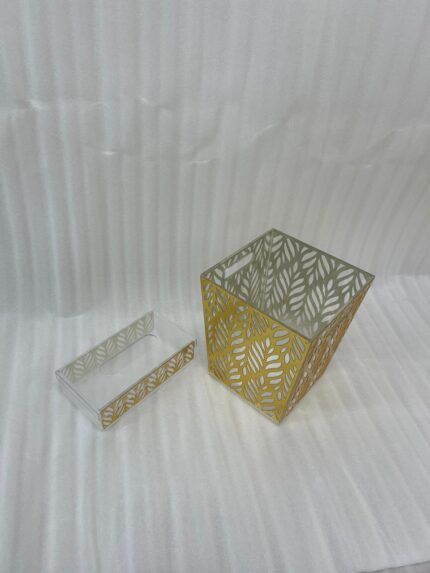 An elegant gold and clear acrylic dust bin with a laser-cut leaf pattern on a wooden table, featuring a matching tissue box holder and a hand dropping a crumpled tissue inside.