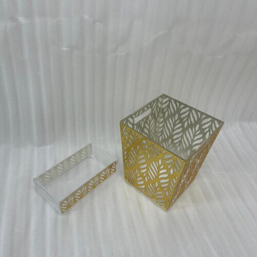 An elegant gold and clear acrylic dust bin with a laser-cut leaf pattern on a wooden table, featuring a matching tissue box holder and a hand dropping a crumpled tissue inside.