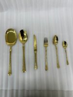 A collection of 36 gold-finished hammered cutlery pieces, including forks, knives, and various spoons, arranged neatly on a grey linen cloth beside a modern kitchen sink.