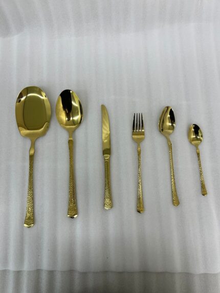 A collection of 36 gold-finished hammered cutlery pieces, including forks, knives, and various spoons, arranged neatly on a grey linen cloth beside a modern kitchen sink.