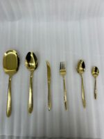 A 36-piece luxury gold cutlery set arranged on a linen cloth next to a kitchen sink, featuring intricate floral engravings on the handles, including assorted dinner spoons, tea spoons, forks, knives, and serving utensils.