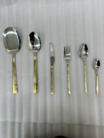 An elegant 36-piece stainless steel cutlery set with gold-finished floral engraved handles, arranged neatly on a dark placemat atop a wooden table, consisting of spoons, forks, and knives.