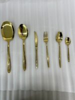 36-piece gold-finished cutlery set featuring delicate floral engraving on handles, arranged on a linen napkin, including table spoons, tea spoons, forks, knives, and serving utensils.