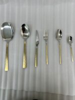 Six pieces from a 36-piece premium cutlery set featuring polished stainless steel heads and intricate, decorative gold-tone handles, displayed on a grey fabric surface near a modern kitchen sink.