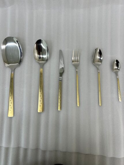 Six pieces from a 36-piece premium cutlery set featuring polished stainless steel heads and intricate, decorative gold-tone handles, displayed on a grey fabric surface near a modern kitchen sink.