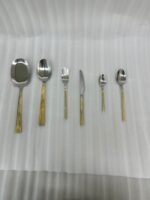 Flat lay of a 36-piece stainless steel cutlery set with polished silver heads and intricately engraved gold floral-patterned handles, arranged neatly on a textured beige linen cloth.