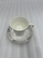 Elegant white Super Bone China teacup and saucer featuring a grey floral pattern and gold rim, placed on a wooden table next to a silver spoon and lavender vase