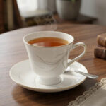 Steaming hot tea in an elegant white bone china teacup with gold trim and a silver leaf design, resting on a matching saucer on a wooden table