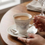 A person holding a white bone china teacup filled with milk tea, featuring a grey floral design and gold rim, resting on a matching saucer with a silver spoon on a wooden table.